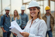 © Vera - Confident Female Architect Smiling at Construction Site with Blueprint, Surrounded by Project Team.
