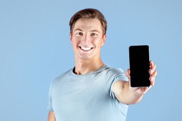 Happy young sportsman showing smartphone with empty screen over lilac studio background, mockup for mobile app design. Strong millennial guy holding cellphone with space for website template