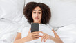 © Prostock-studio - Amazing or bad news, strange offer during covid-19 lockdown. Shocked young zoomer african american female lies in soft bed and looks at smartphone, reading message in bedroom, top view, free space