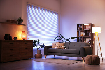 Poster - Stylish living room with comfortable sofa and wooden shelving unit in evening. Interior design