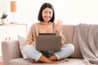 © Prostock-studio - Online Video Call. Portrait of smiling asian lady making virtual conference on laptop, waving with hand to webcam. Communication Concept. Woman chatting using computer, sitting on the couch at home