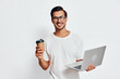 © SHOTPRIME STUDIO - Happy cheerful young man holding a laptop in one hand and a coffee cup in the other, smiling at the camera against a plain white background. Work lifestyle concept.