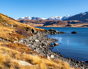 Wall Mural - serene mountain lake with rocky shore, surrounded by golden grass and distant snow capped peaks, evokes sense of tranquility and natural beauty