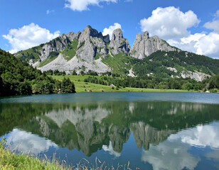Wall Mural - serene landscape featuring towering jagged mountains reflected calm lake, surrounded by lush greenery and clear blue sky with fluffy clouds. scene evokes sense of tranquility and natural beauty