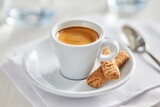 A white cup of espresso on a saucer with biscotti biscuits, next to a spoon, on a light table setting.