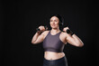 © New Africa - Plus size woman in gym clothes with headphones and dumbbells on black background