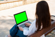 © timoha.ig - Young caucasian female using laptop with green screen outdoors on a sunny day, sitting on a wooden bench in casual attire, rear view. Digital nomad lifestyle. Remote work
