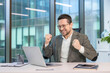 © Tetiana - Happy young businessman sitting in the office at his desk, looking at the laptop screen and rejoicing, showing a gesture of victory and success