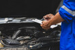 © Wasan - Car mechanic inspecting engine and taking notes on clipboard. Mechanic in a uniform is holding a clipboard and inspecting a car engine, taking notes while assessing the vehicle's condition.