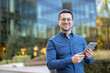 © Liubomir - A smiling man, wearing glasses and a blue shirt, holds a tablet in front of a modern building.