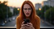 © alleb - Woman with red hair looks shocked while holding a smartphone outdoors.