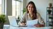 © Nathan - Smiling young woman of Caucasian descent sitting at a desk with laptop, reviewing documents in a bright office setting.