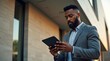 © Nathan - A young Black man in a suit focuses on his tablet, standing outside a modern building in soft evening light.