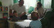 © Marco - Toddler leaning on sofa watching family interaction, father playing train with boy at coffee table, mother smiling nearby, intimate family bonding moment indoors
