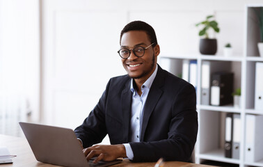 Cheerful african american businessman working with modern laptop in office, copy space. Happy young black manager in suit and glasses working online, typing emails or chatting with clients