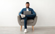 © Prostock-studio - Young Arab man in casual wear working online, sitting in armchair and using laptop against white studio wall, full length. Millennial Eastern guy surfing internet on portable pc