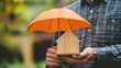 © ARIF - An insurance agent is holding a orange umbrella over a wood house model. Property insurance concept.
