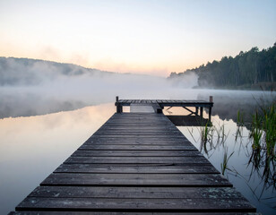 Wall Mural - serene wooden pier extending into misty lake at dawn, surrounded by lush greenery and soft fog. tranquil atmosphere evokes sense of peace and calmness