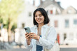 © Andrei - Waiting for directions to load, a stylish young woman of mixed heritage scrolls her phone against a charming European backdrop