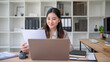 © wattana - Young professional woman reviewing paperwork with a laptop in a clean, organized workspace.