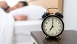 © intricateflow - A traditional analog alarm clock on a nightstand facing the camera, softly lit, with a peaceful background of a person still sleeping in bed, capturing a serene early morning bedroom scene