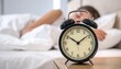 © intricateflow - A traditional analog alarm clock on a nightstand facing the camera, softly lit, with a peaceful background of a person still sleeping in bed, capturing a serene early morning bedroom scene