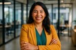 © Velar - In an office setting, a smiling Latin American businesswoman stands confidently with her arms crossed, facing the camera