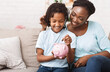 © Prostock-studio - Money Saving Concept. Happy black mom and daughter inserting coins in pink piggy bank, empty space