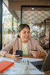 © Satori Studio - Smiling woman using smartphone and credit card for online shopping in a cafe, with documents and laptop on the table.