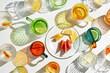 © yrabota - Summer still life scene with colored glass cups with handles and plate with slices of fruit. Glittering glasses of water on white table at sunlight. Long colors shadows. Summer concept, above