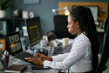 African American woman diligently working at her desk, analyzing data on computer screen while sitting in a modern office setting. She appears focused and engaged