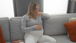 © Krakenimages.com - Pregnant woman with blonde hair sitting on a sofa in a cozy living room touching her belly, conveying a sense of calm and expectation in her home environment.
