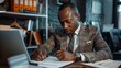 © Shankar - African american man in suit writing on paper at desk with laptop and office supplies in background