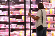 © Andriy Medvediuk - Rear view of adult female customer opens big glass door in supermarket's fridge and chooses packaged fresh chilled meat. Assortment of meat on counter of hypermarket. Meat showcase in market place.