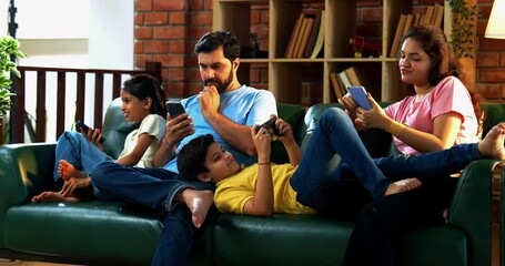 Indian family using smartphones individually while sitting together on living room couch, showing screen addiction in modern lifestyle, no interaction or bonding, lack of quality family time concept