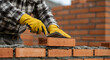 © abu - Construction Worker Carefully Laying Red Bricks with Trowel and Mortar Wearing Yellow Gloves on a Cloudy Day