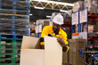 © Rakchanok - Warehouse packer applies fragile label onto box at packing station, ensuring careful handling before freight leaves bustling distribution center with shelves of goods. African male, safe posture
