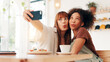 © peopleimages.com - Selfie, tongue out and women in coffee shop, silly and photography for memory of reunion or embrace. Goofy, friends and bonding on break with profile picture, happy or social media on weekend in cafe