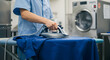 © abu - Close Up of Person Ironing Blue Garment with Steam in Laundry Room with Washing Machines in Background