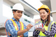 © NANKHAMMAI - Male and female engineers wearing hard hats smile and discuss blueprints on a construction site, demonstrating teamwork, safety and expertise.