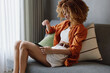 © SHOTPRIME STUDIO - Young woman using a tablet at home, wearing a stylish orange shirt and white shorts, expressing calmness in a cozy interior with soft colors