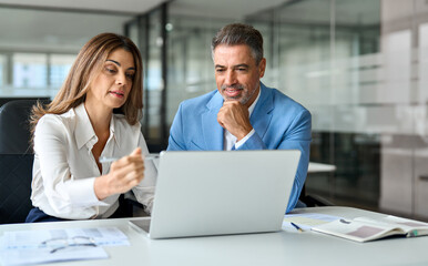 Fototapete - Two executive leaders team using laptop working on computer at work desk, busy happy middle aged professional business man and woman having conversation on financial project at meeting in office.