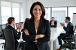 © Alex & M Studio - Confident businesswoman with arms crossed smiling in modern corporate office environment. Professional female executive in formal attire with blurred colleagues in background