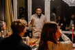 © Drobot Dean - A Black adult man stands and makes a toast with a glass of wine while multiracial adults sit around a festive dinner table in a modern home at night.