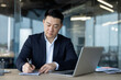 © Tetiana - An Asian businessman diligently taking notes while working on his laptop in a modern office setting.