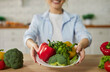 © Studio Romantic - Cropped shot of smiling woman showing plate with fresh vegetables including bell peppers, broccoli and herbs at camera in modern kitchen. Healthy eating, vegetarian diet and organic food concept.