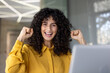 © Liubomir - A joyful woman with curly hair celebrates success, raising her fists with an excited expression, in a bright setting.
