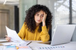 © Liubomir - A woman with curly hair, wearing a yellow shirt, reviews documents, looking tired while working on her laptop in the office setting.