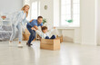 © Studio Romantic - Happy family, new flat home owners, parents playing with cute small kid son on moving day, tenants, renters homeowners and child having fun riding in cardboard box in living room, relocating