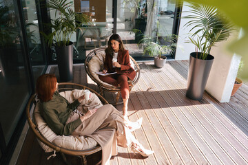 Fototapete - Professional ladies having conversation sharing ideas using tab at work. Two busy business women of young and middle age talking in green cozy office sitting in chairs. Candid shot. Top view.
